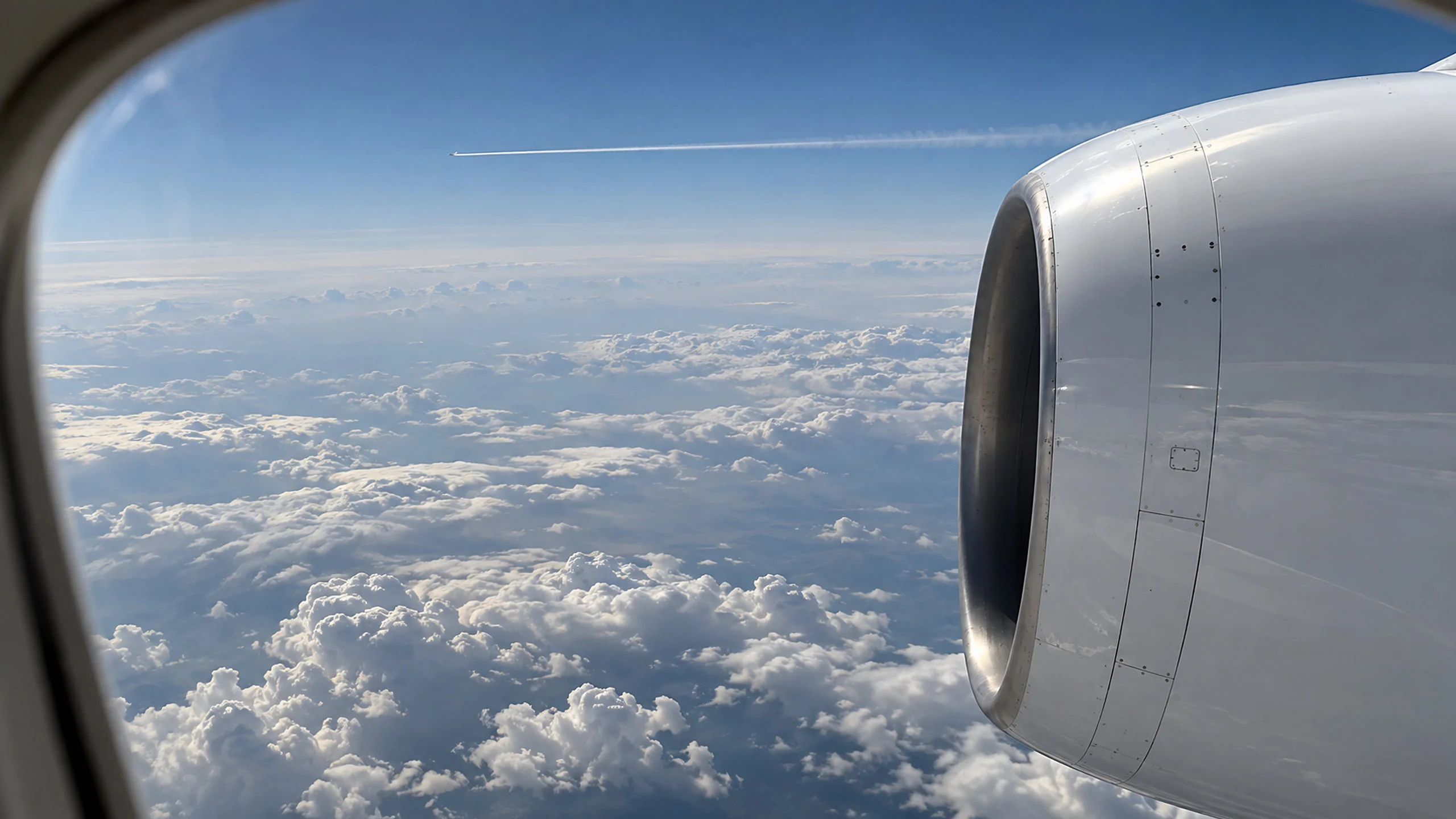 View from a jet's cabin window at cruise altitude: the engine nacelle beside a deep cloudscape, and the thin contrail of a distant aircraft in the blue above