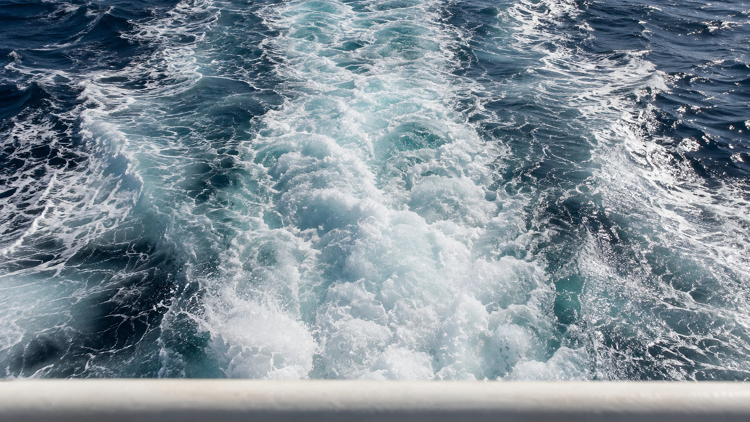 The white wake of a ship at speed, seen from the stern rail, foam streaming away across dark-blue water